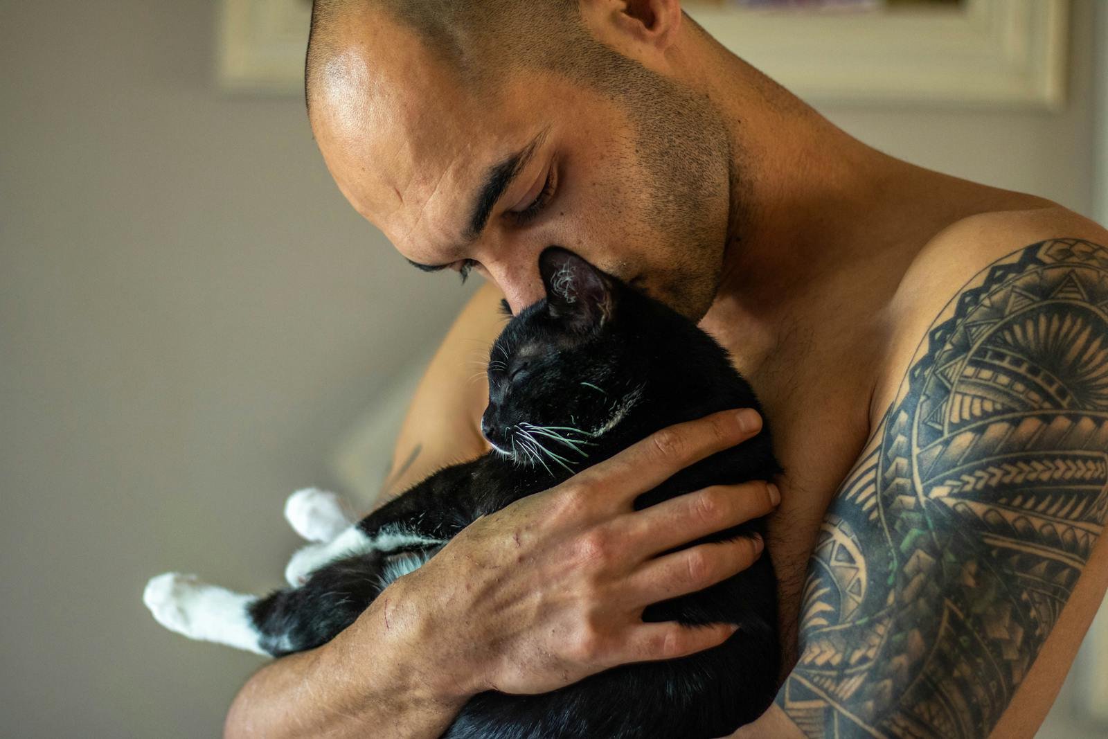 Shirtless man lovingly holding a black cat indoors. Tattoo visible.