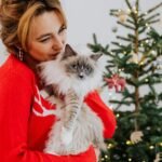 A woman in a red sweater cuddling her fluffy cat near a decorated Christmas tree.