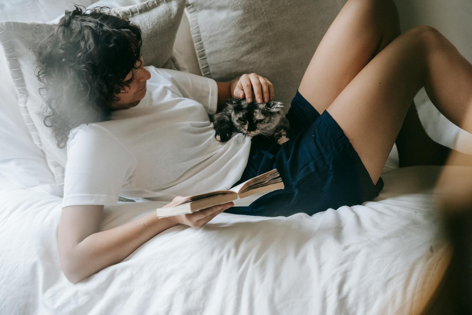Side view content young female in casual clothes stroking cute cat while resting on cozy bed with book