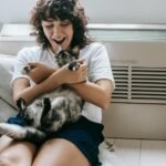 Happy female with dark hair caressing adorable dappled cat while sitting on floor with hands crossed near radiator and bed in room at home