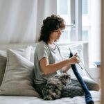 Adorable cat sitting on legs of concentrated young ethnic female owner with curly hair knitting on bed during weekend at home
