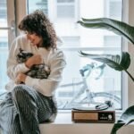 Young woman in cozy attire hugging her cat by the window, surrounded by plants and a retro turntable.