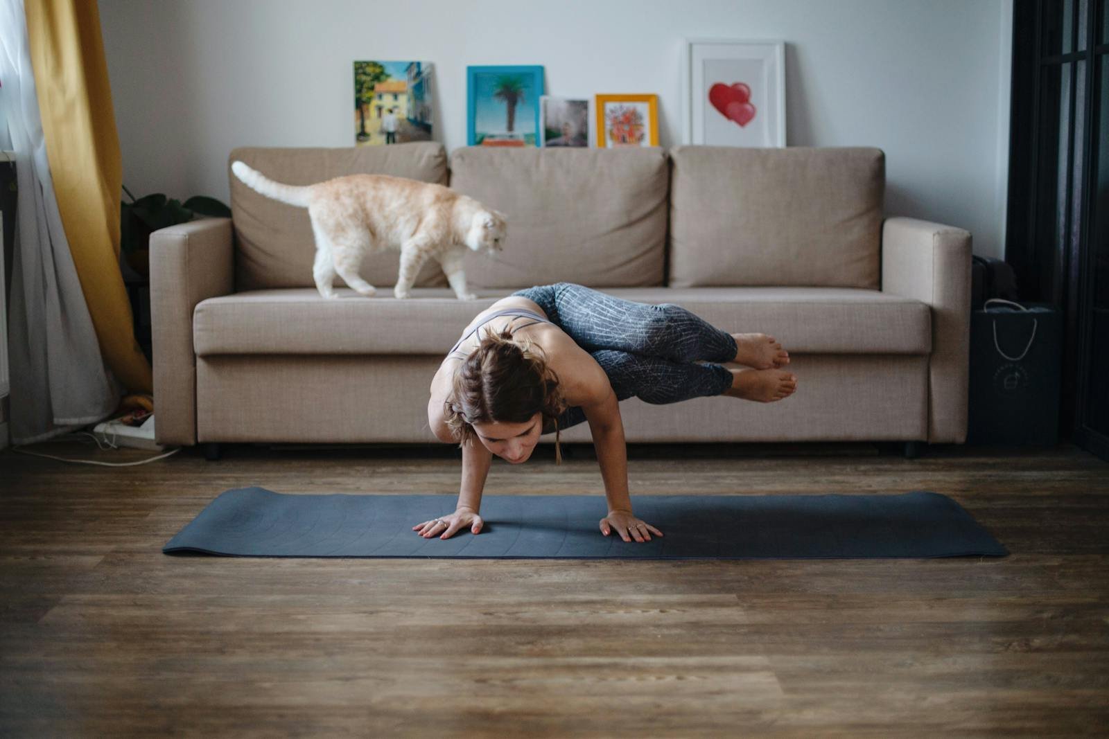 A woman performing a yoga handstand indoors, with a cat walking on the sofa behind her.