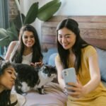 Three women smiling and taking a selfie with a cat in a cozy bedroom setting.