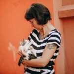 Woman with tattoos gently holding a cat against an orange wall.