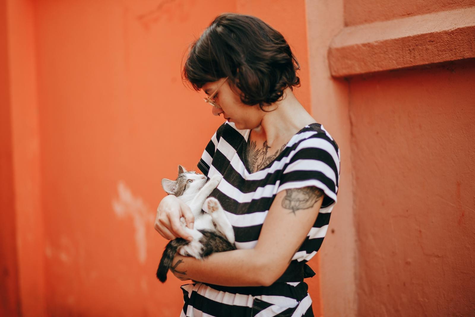 Woman with tattoos gently holding a cat against an orange wall.