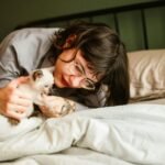 A woman lovingly playing with her Siamese kitten on a bed.