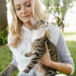 A woman sits outdoors holding her tabby cat in a sunny park setting.
