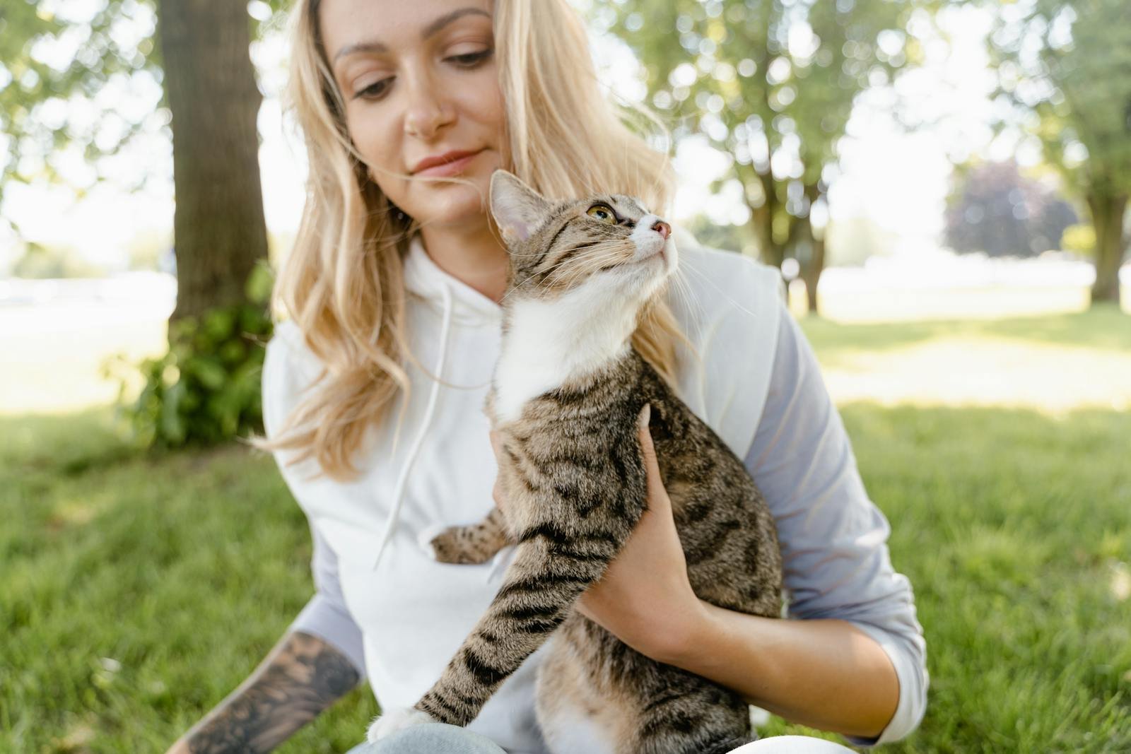 A woman sits outdoors holding her tabby cat in a sunny park setting.