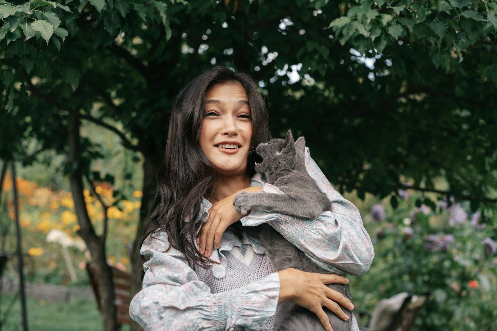 A joyful woman holding a cat in a vibrant garden captures a moment of playful affection.