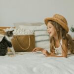 Joyful child in a straw hat playing with a cat on a bed, surrounded by cozy decor and plants.