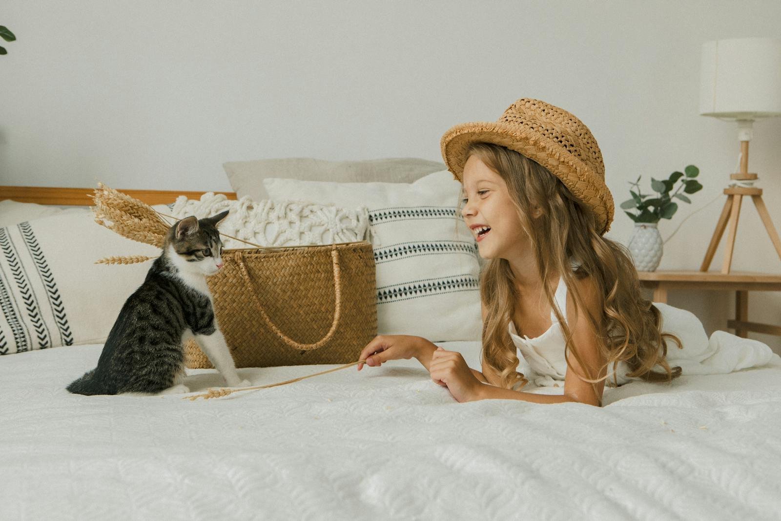 Joyful child in a straw hat playing with a cat on a bed, surrounded by cozy decor and plants.