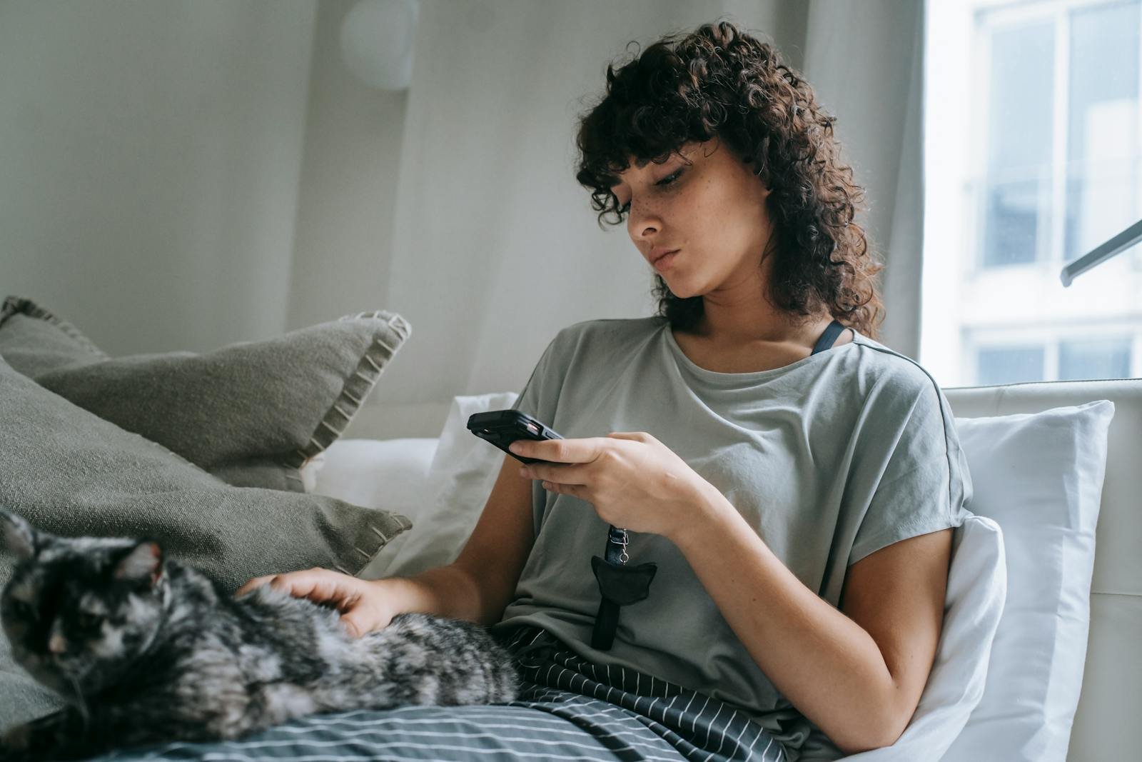 Woman sitting on a bed with a cat, using a smartphone, enjoying a calm, cozy moment indoors.