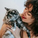 Crop cheerful female with red lips and freckled skin embracing adorable cat with spotted fur in daytime